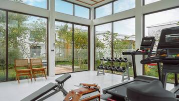 A spacious gym with large windows, a treadmill, a rowing machine, dumbbells, and two wooden chairs.