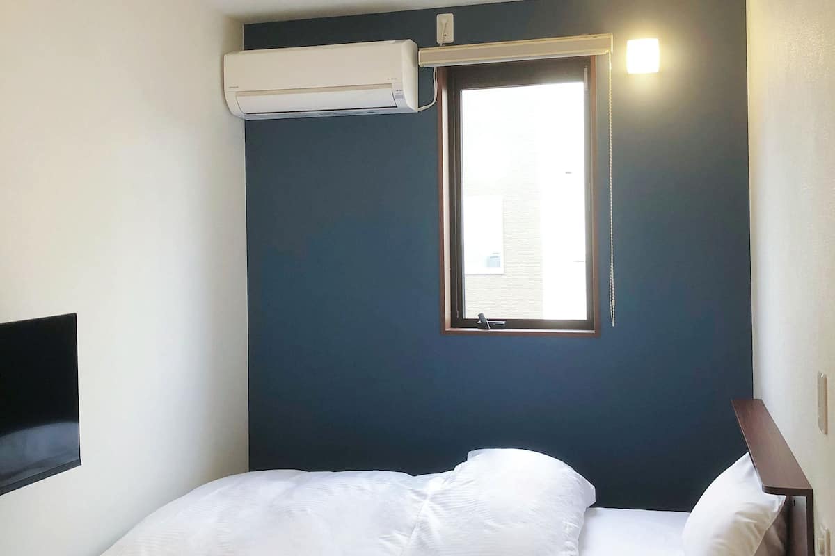 Een eenpersoonsbed met wit beddengoed, een klein nachtkastje, een raam met uitzicht en een airconditioningunit aan de muur.