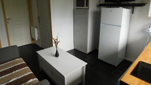 A compact living space with a kitchenette, a flat-screen TV mounted above the fridge, a clock on the wall, and a small table with a vase. - Short-term furnished studio rental (n° 4) (Le Trait)