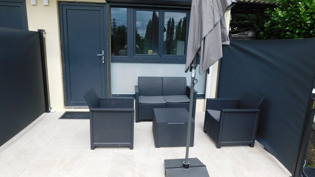 A patio area with a table and four chairs, a folding umbrella, and a door with a window. - Short-term furnished studio rental (n° 4) (Le Trait)