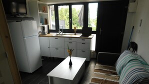 A compact kitchen with a white countertop, a sink, and a dining table with a vase of flowers. There is a sofa with striped cushions and a window with a view of greenery. - Short-term furnished studio rental (n° 4) (Le Trait)