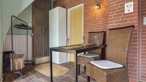 Una habitación con pared de ladrillo, puerta de madera, una mesa con sillas, una silla de mimbre y un perchero.