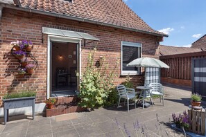 Et murstenshus med tegltag, en terrasse med bord og stole samt plantekrukker.