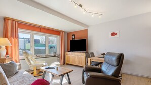 A living room with a sofa, armchair, dining table, and a TV on a cabinet. - Apartment 'Ferienwohnung Kapitänskajüte' with Private Terrace, Shared Garden and Wi-Fi (Nordstrand)