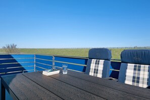 Ein Tisch mit zwei Stühlen, einem Glas Wasser und einem Buch auf einer Terrasse mit blauer Reling und Blick auf ein Feld.