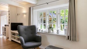 Ein Wohnzimmer mit einem grauen Sessel, einem kleinen Hocker, einem Fernseher auf einem weißen Schrank, einem Fenster mit Vorhängen und einer weißen Wand mit einem Regal.