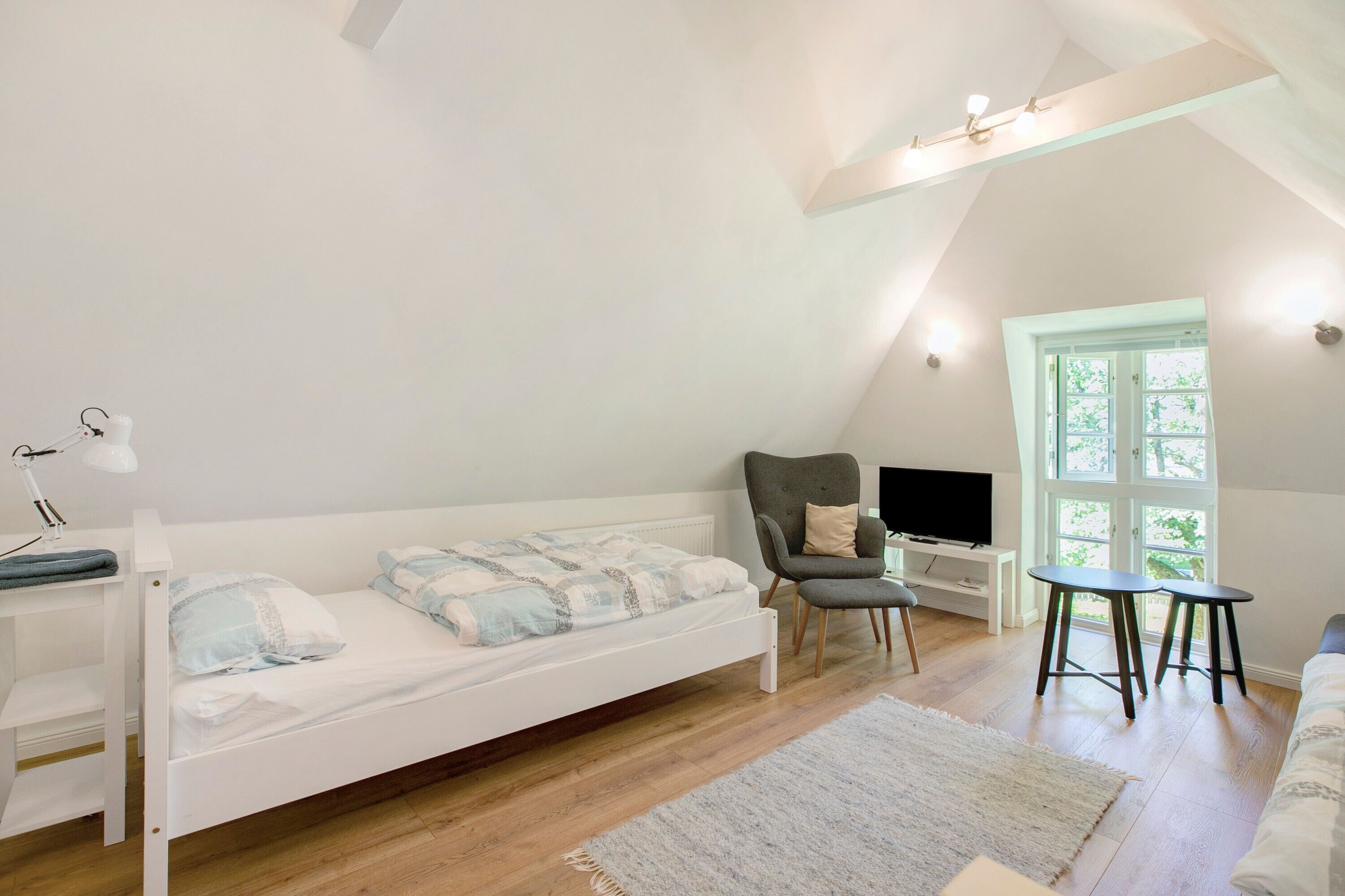 A bedroom with a bunk bed, a desk with a lamp, a chair, a TV, and a window with a view of greenery.