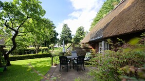 Ein Haus mit Strohdach, eine Terrasse mit Tisch und Stühlen, umgeben von viel Grün.