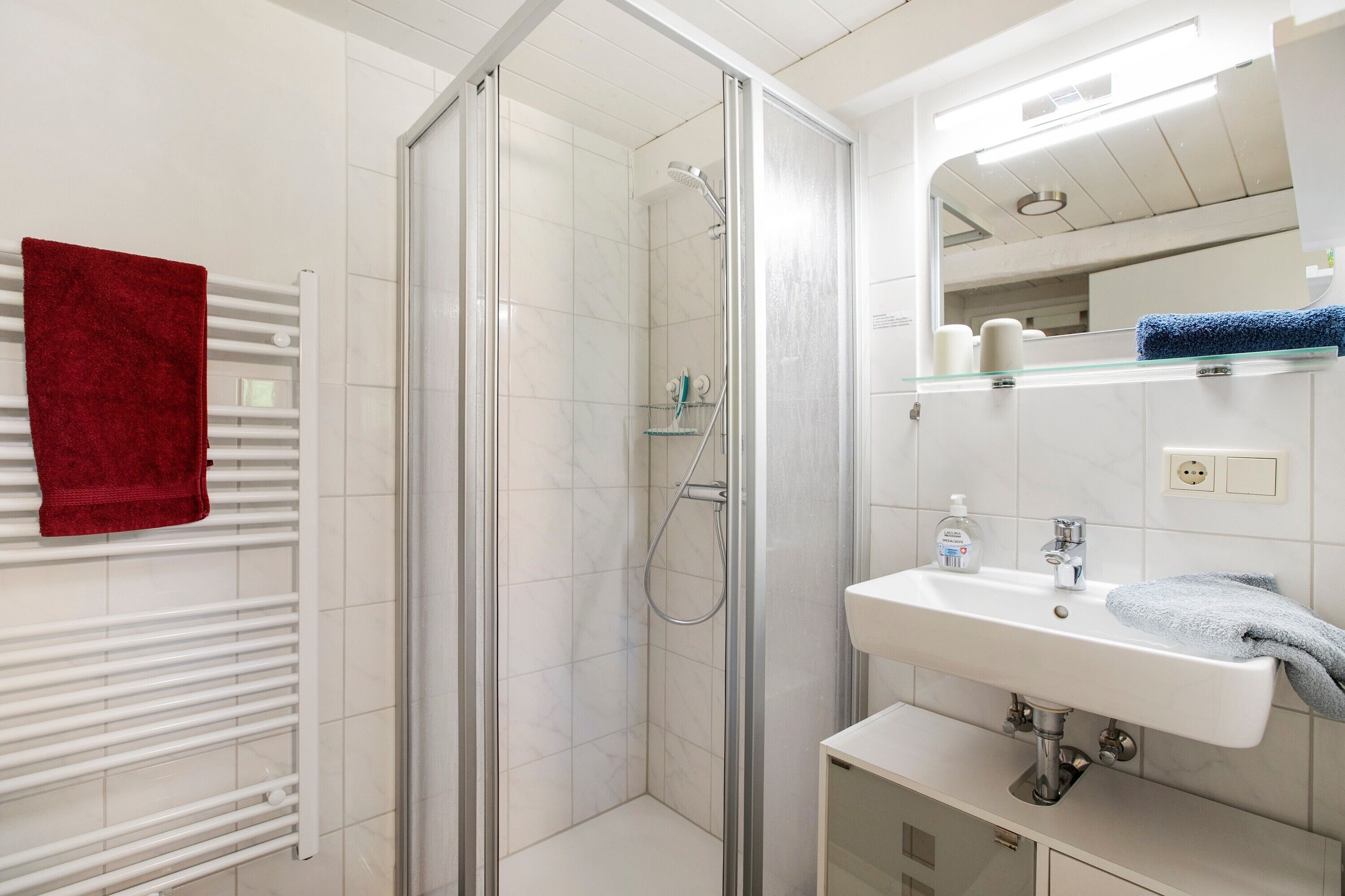 Une salle de bain avec une douche à parois de verre, un lavabo blanc et une serviette rouge posée sur un radiateur.