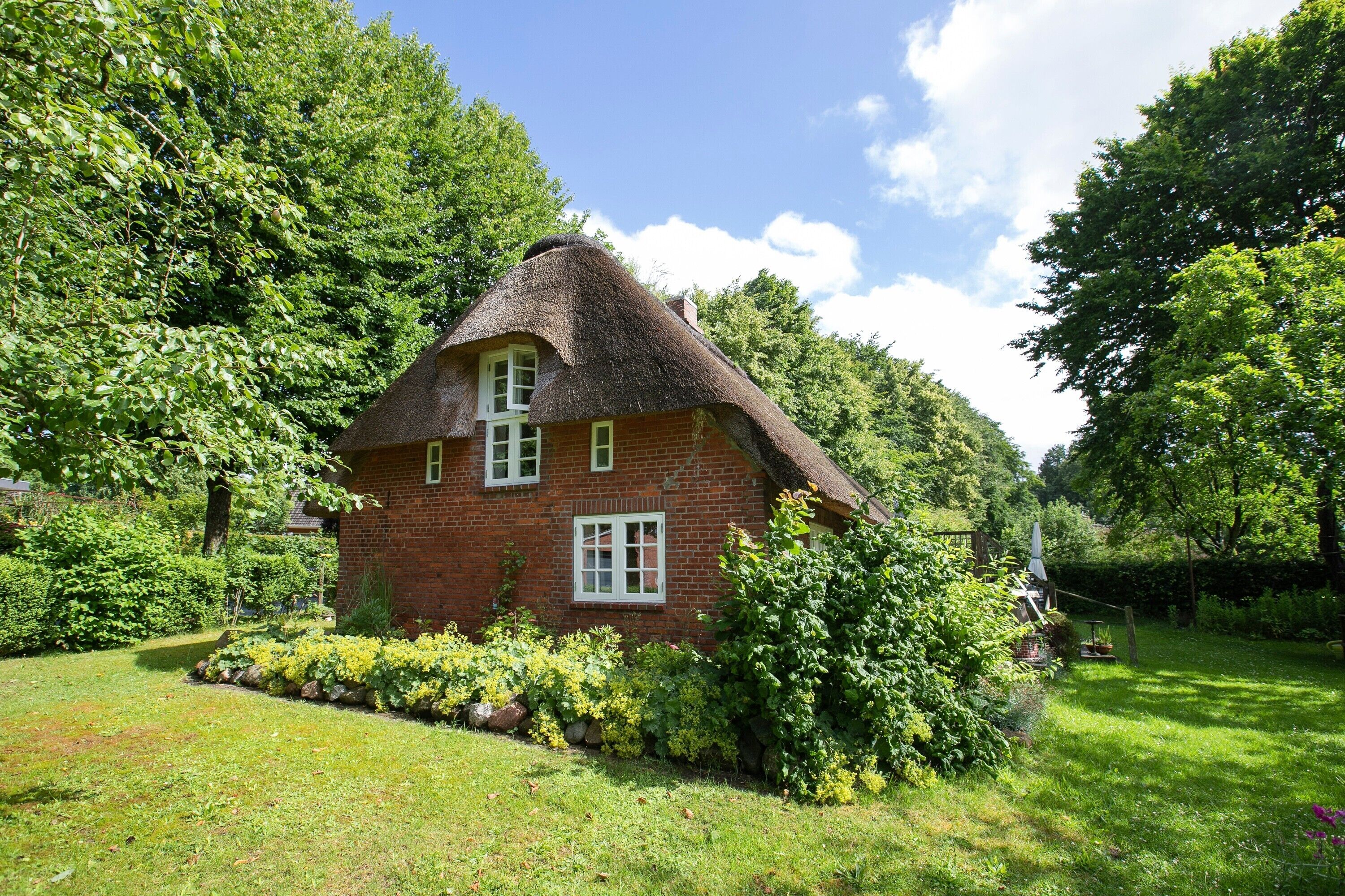 A red brick house with a thatched roof, surrounded by lush greenery and a well-maintained garden.