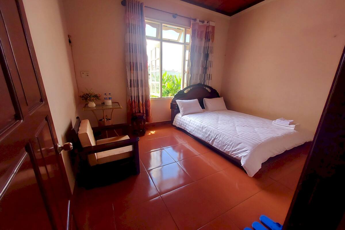 A bedroom with a bed, a chair, a small table with a lamp, and a window with curtains.