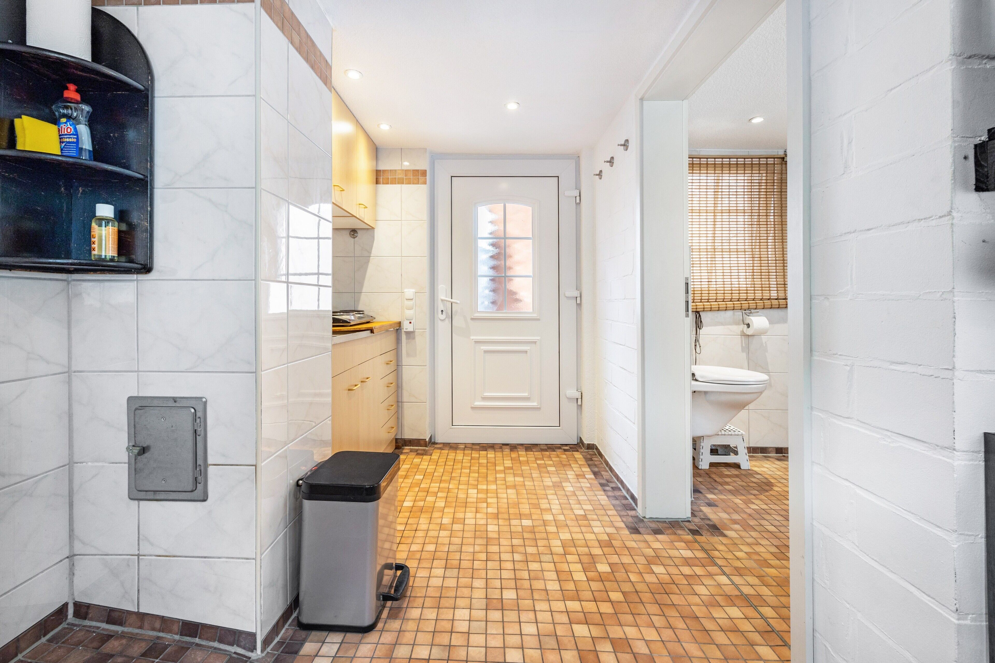 Un couloir étroit avec un sol carrelé, une porte donnant sur une salle de bain et une poubelle.