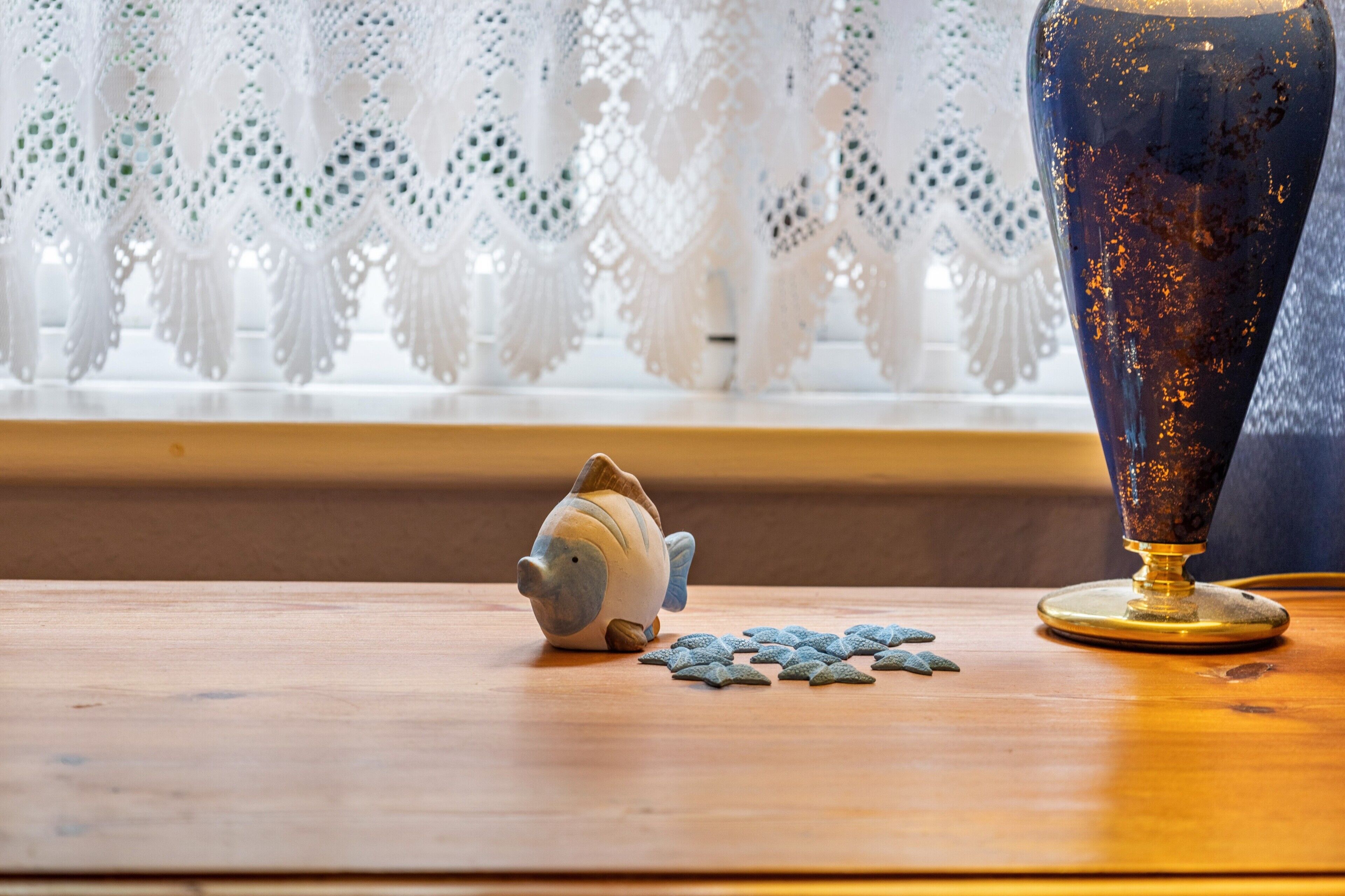 Una mesa de madera con un pez y una estrella de mar de cerámica, un florero y cortinas de encaje al fondo.