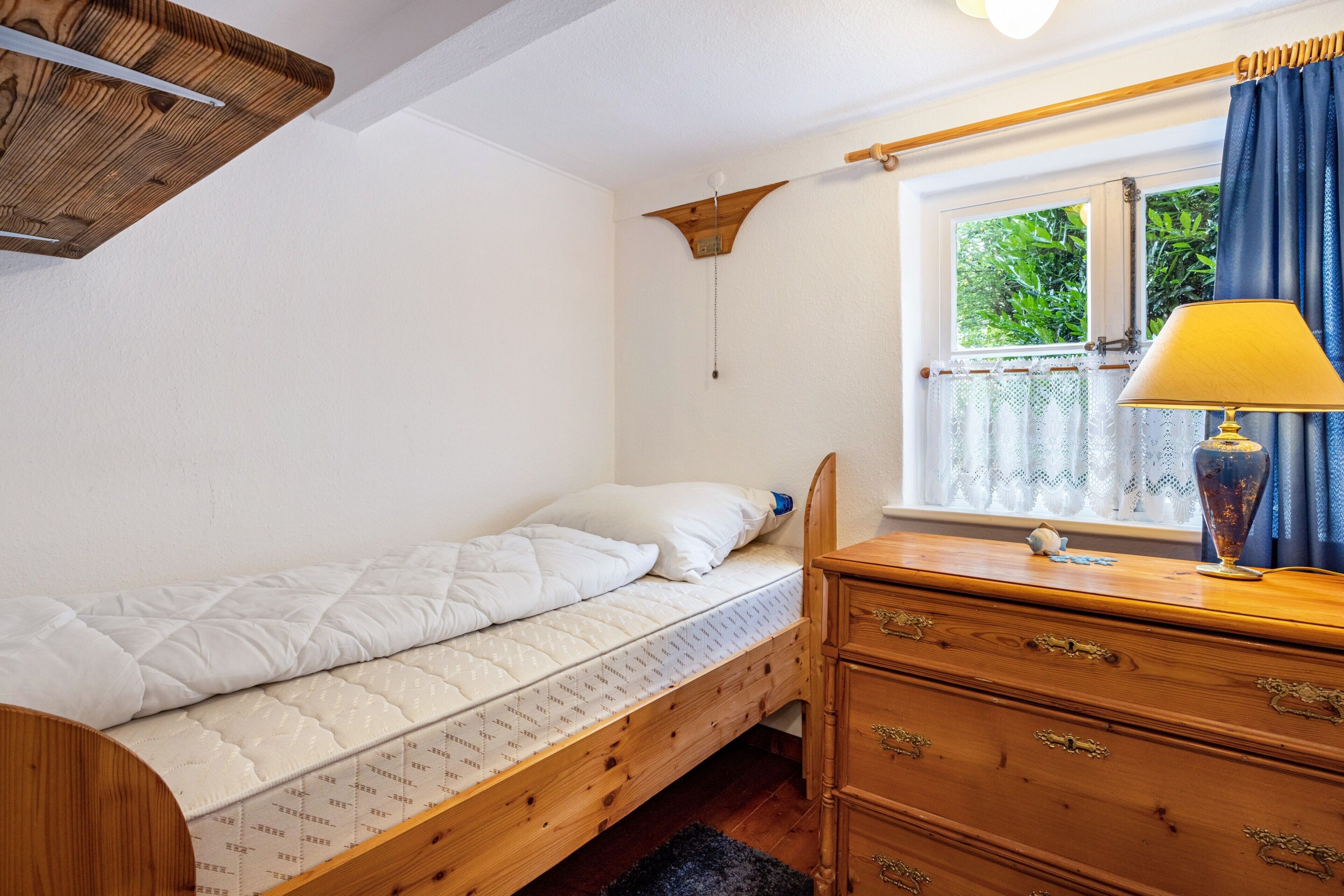 Una cama individual con cabecera de madera, un aparador de madera con cajones, una mesita de noche con lámpara y una ventana con cortinas.