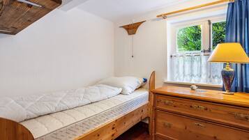 Una cama individual con cabecera de madera, un aparador de madera con cajones, una mesita de noche con lámpara y una ventana con cortinas.