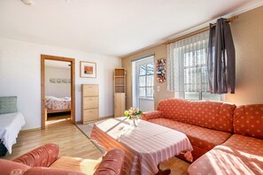 Una habitación con sofá, una mesita con un florero, cama, armario y ventana con cortinas.
