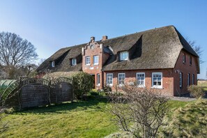 Une grande maison en briques avec un toit de chaume, une petite dépendance attenante et un jardin clôturé.
