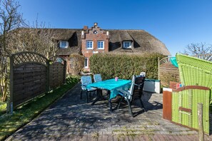 Une maison en briques avec un toit de chaume, une terrasse dallée avec une table et des chaises, et un portail en bois.