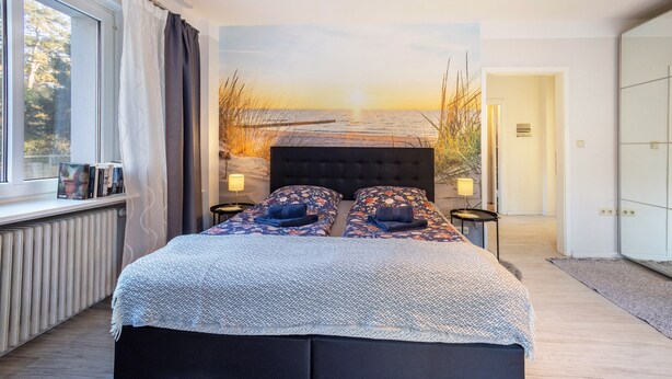 A bedroom with a large bed, a view of the sea through the window, a radiator, and a wardrobe. - Holiday Home 'Sandstrand' with Private Terrace, Garden, and Wi-Fi (Timmendorfer Strand)