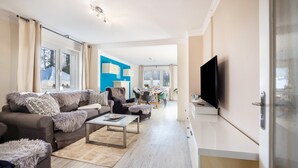 A modern living room with a sofa, coffee table, and TV, leading to a dining area with chairs and a table. - Holiday Home 'Sandstrand' with Private Terrace, Garden, and Wi-Fi (Timmendorfer Strand)