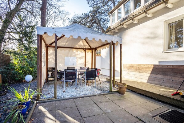 Un patio couvert, équipé d’une table et de chaises, entouré d’une terrasse en bois et d’un sol en gravier.
