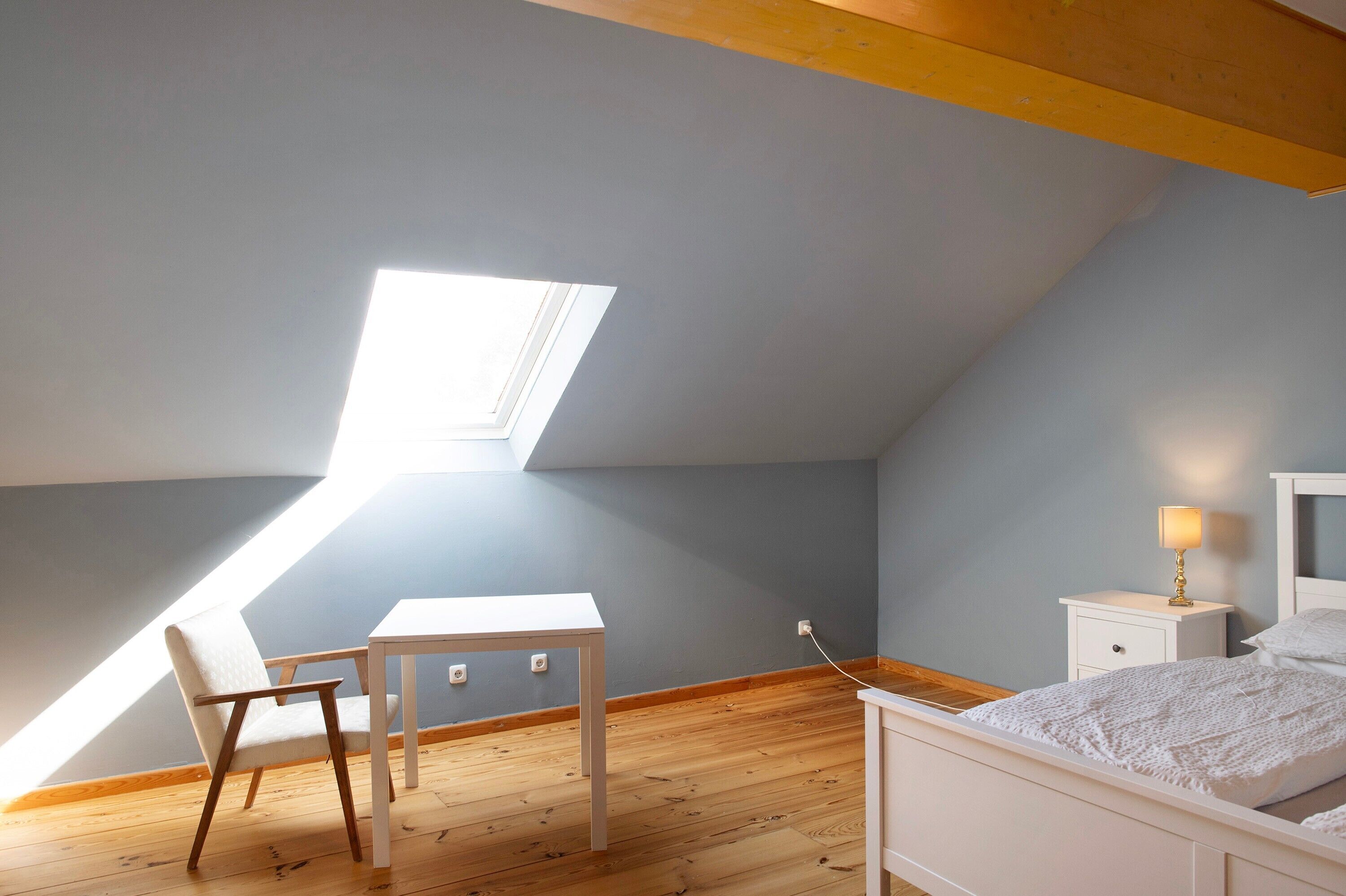 Ein Schlafzimmer mit schrägen Wänden, einem Oberlicht, einem Bett, einem Nachttisch, einem Sessel und einem kleinen Tisch.