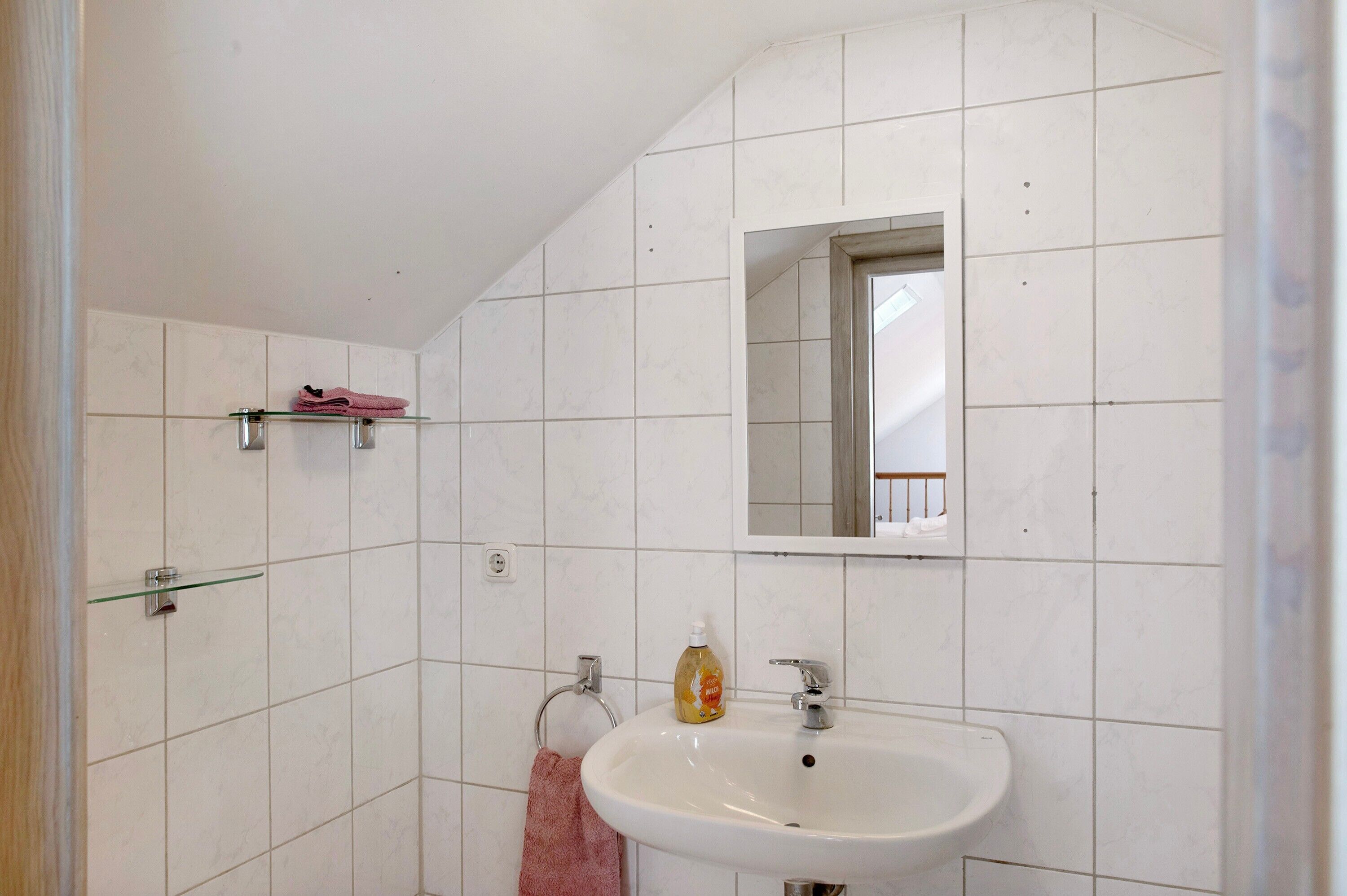 A bathroom with white tiles, a sink, a mirror, and a glass shelf.