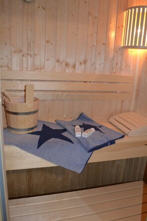 Een houten sauna met een bank, een houten emmer, een handdoek en een wandlamp.