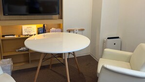 Una habitación pequeña con una mesa redonda blanca, una silla de cuero blanco, un televisor montado en la pared y un estante con diversos objetos, incluyendo un reloj, un pulverizador y un control remoto.