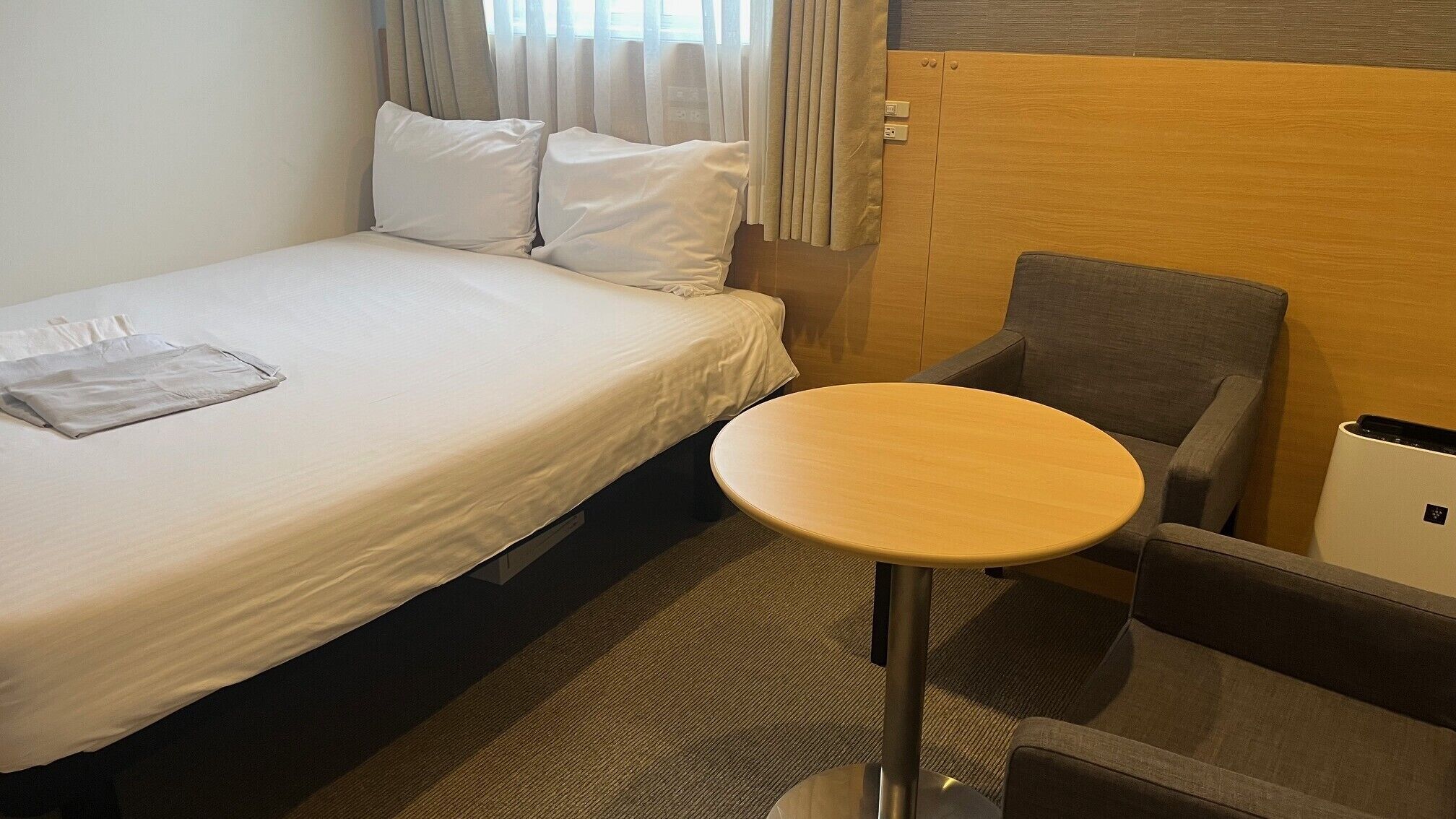 A hotel room with a bed, a small round table, a chair, and a trash bin.