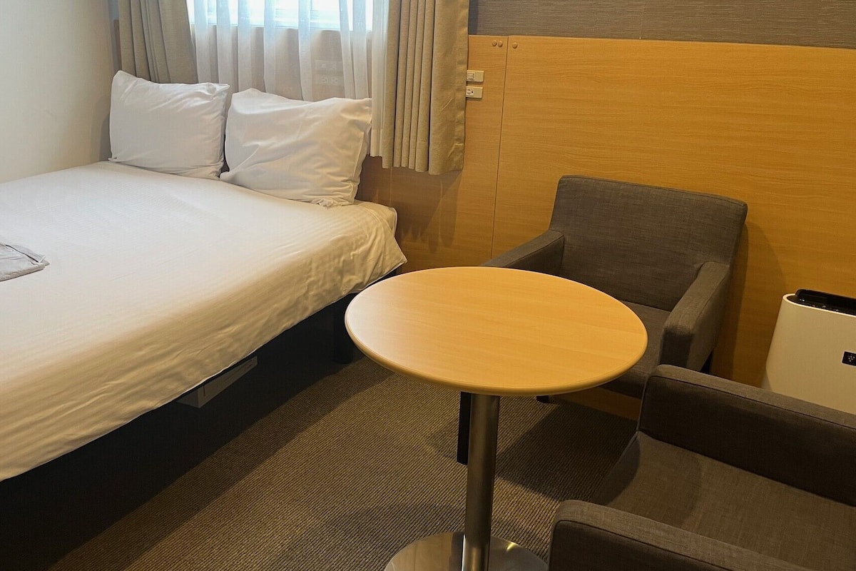 A hotel room with a bed, a small round table, a chair, and a trash bin.