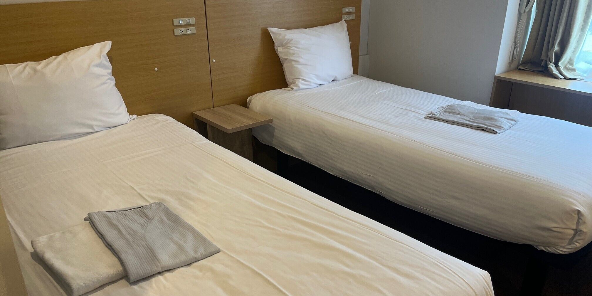 Dos camas individuales con sábanas y almohadas blancas, un cabecero de madera, una mesita de noche y una ventana con cortinas.