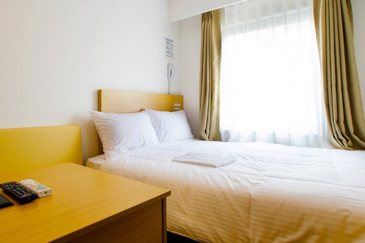 A hotel room with a bed, a bedside table with a remote control, and a window with curtains.