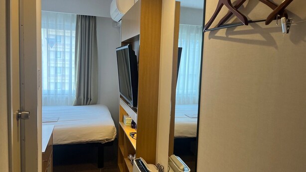 Una habitación de hotel compacta con cama, televisor, escritorio con ordenador y ventana con cortinas.