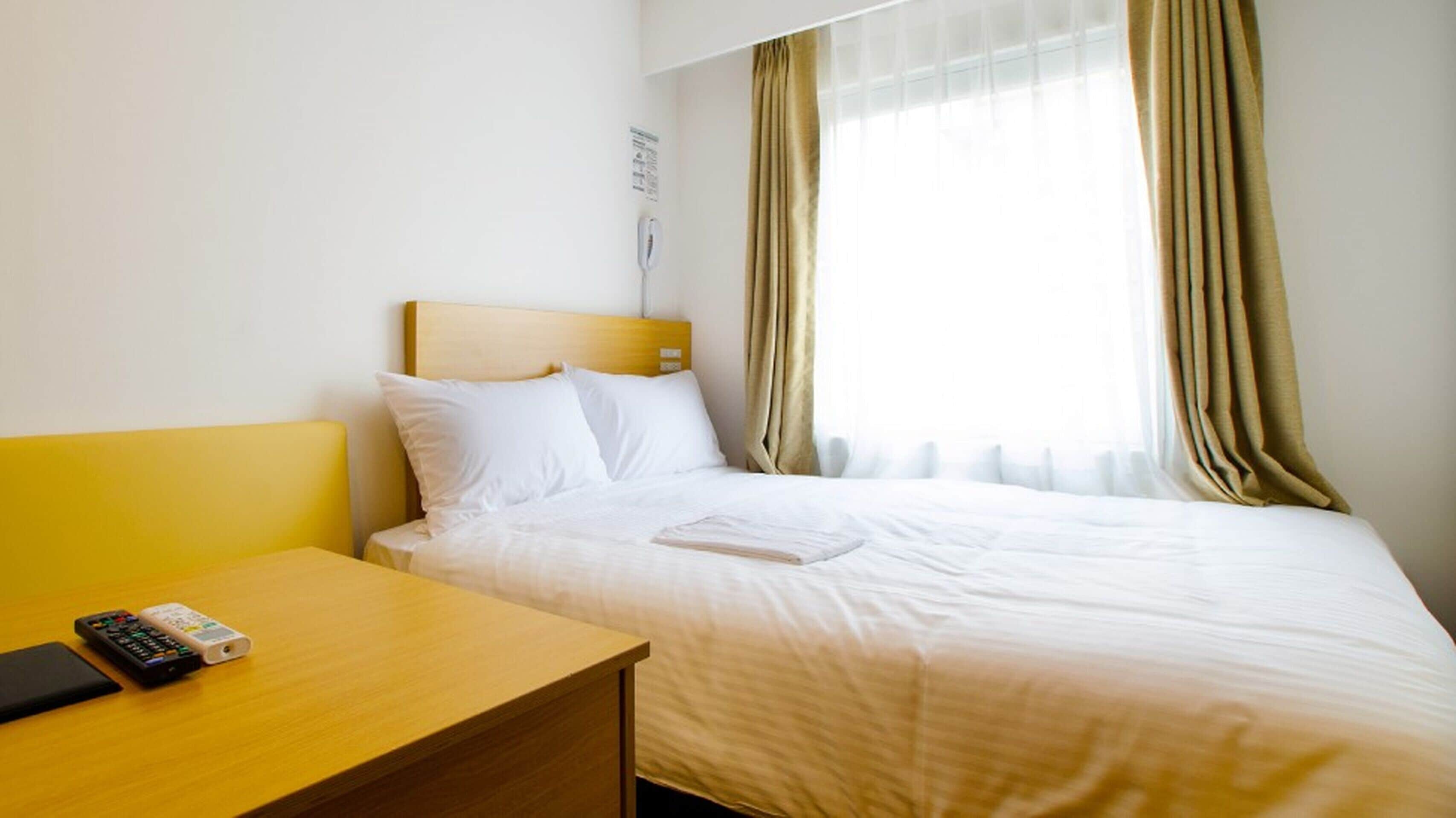 A hotel room with a bed, a bedside table with a remote control, and a window with curtains.