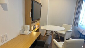 Habitación de hotel con un escritorio de madera, una mesa redonda, dos sillones blancos, un televisor montado en la pared y una cama con edredón azul.