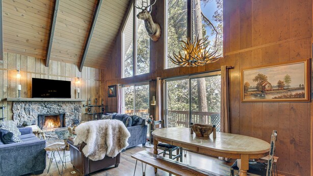 A rustic interior with a fireplace, wooden walls, a dining table, and a large window overlooking nature. - Chic Lake Arrowhead Retreat: 4 Seasons of Fun! (Lake Arrowhead)