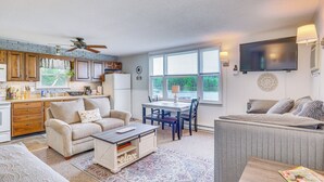 A spacious living area with a sofa, coffee table, and a dining table with chairs. There is a kitchen area with cabinets and a refrigerator. A ceiling fan is visible, and a flat-screen TV is mounted on the wall. - Lake of the Ozarks Cabin w/ Private Dock + Kayaks (Lincoln)