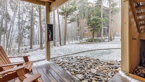 Un patio in legno con un braciere in pietra, una sedia di legno e vista su una zona boschiva con edifici sullo sfondo.