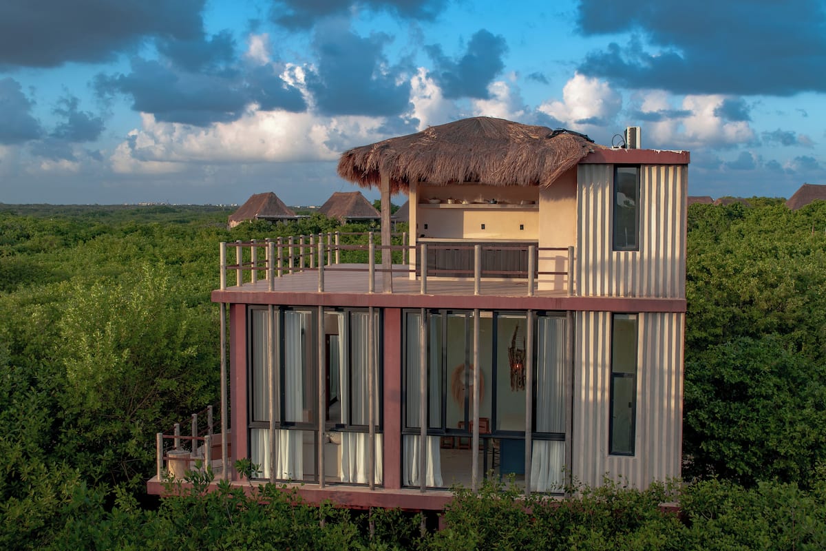 A modern, multi-level building with large windows and a thatched roof structure, surrounded by lush greenery.