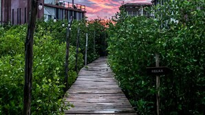 Ein hölzerner Weg führt zu einem Gebäude, an dem ein 'RELAX'-Schild angebracht ist, umgeben von üppiger Vegetation und einem Sonnenuntergangshimmel.