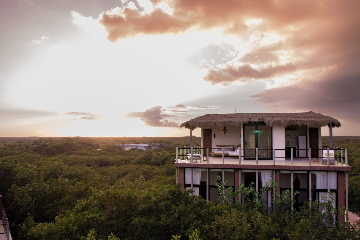 A thatched-roof building with a balcony, surrounded by dense greenery and a sunset sky.