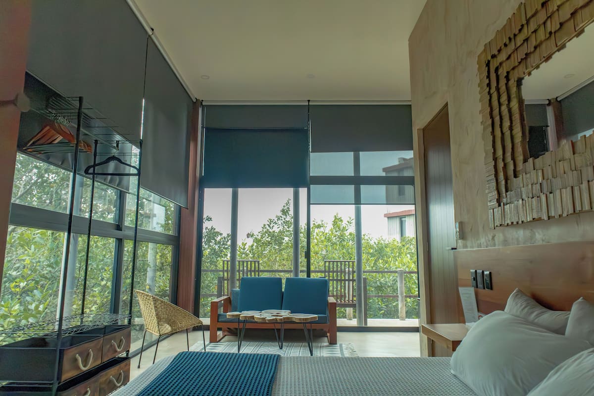 A modern hotel room with a large bed, a sofa, a small table, and a balcony with a view of greenery.