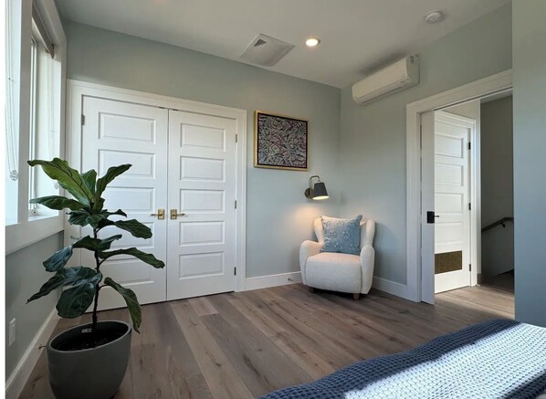 A room with a large potted plant, a white armchair with a patterned cushion, a framed artwork on the wall, and a door leading to another room. - High end home in downtown Tillamook (Tillamook)