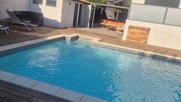 A swimming pool with a wooden deck, lounge chairs, and a covered seating area.
