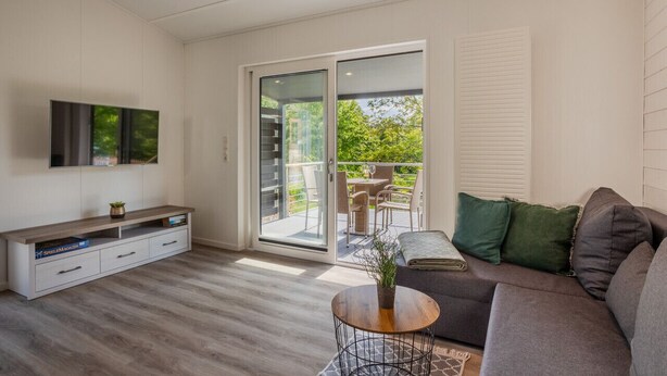 Een moderne woonkamer met een flatscreen televisie, een bank, een salontafel en een glazen schuifdeur die leidt naar een buitenterras met een tafel en stoelen.