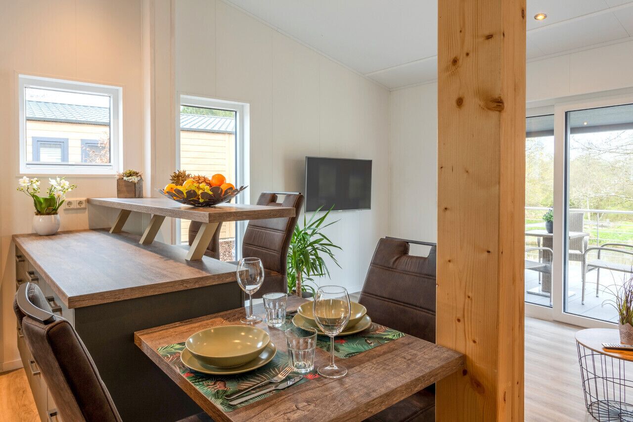 Un coin repas avec une table en bois dressée pour deux personnes, un saladier rempli de fruits et un téléviseur fixé au mur.