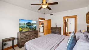 Ein Schlafzimmer mit einem Bett, einem Deckenventilator, einer Holzkommode, einem Fernseher und einer Tür zu einem anderen Raum.