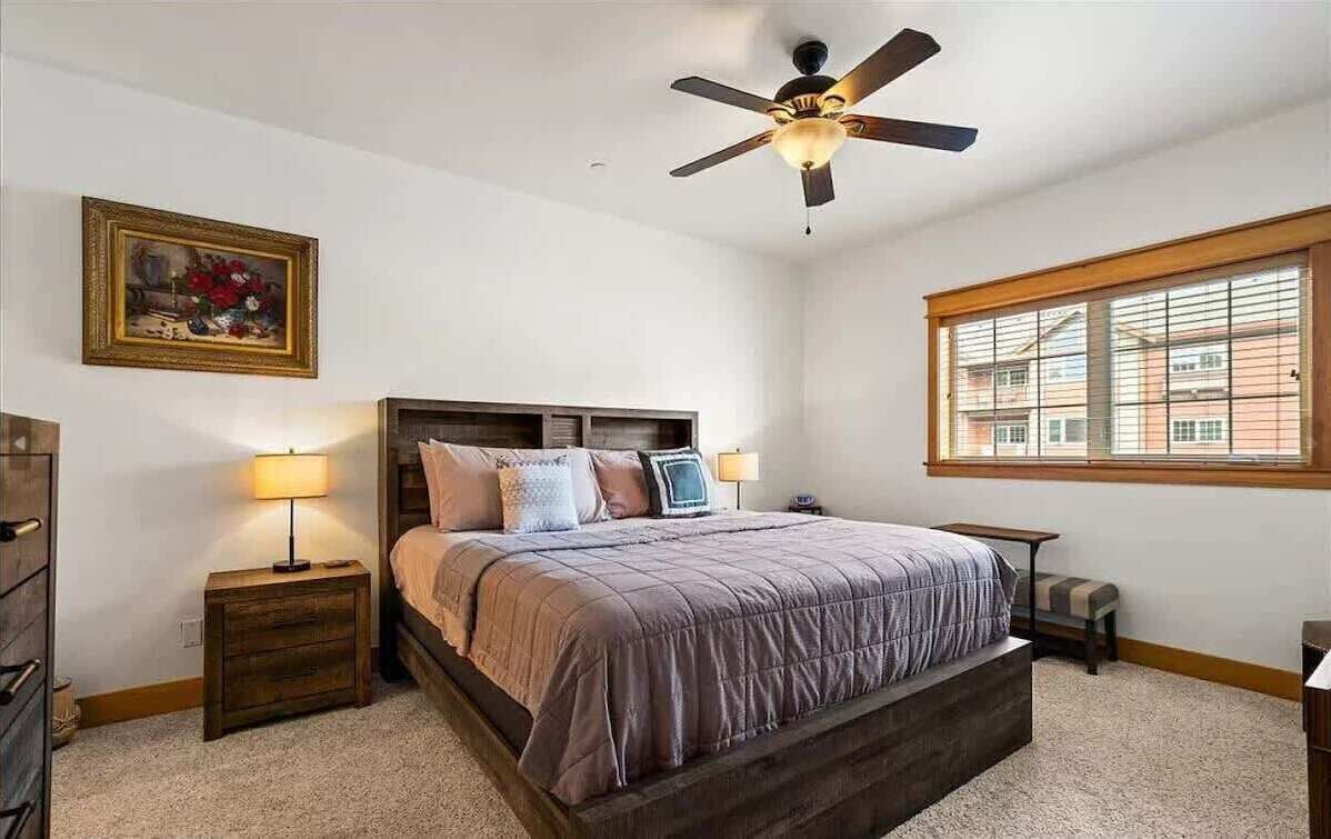 A bedroom with a large bed, two bedside tables, a ceiling fan, a window with blinds, and a framed picture on the wall.
