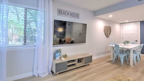 A modern living room with a TV, a dining table with chairs, a sign reading 'BEACH HOUSE', and a large window with blinds. - Steps to Beach | Pool | Pet Friendly | Sleeps 8 | Hilton Head (Hilton Head Island)